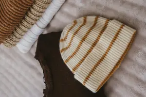 Bonnet en tricot crème à rayures beige, posé sur des vêtements pliés sur une couverture.