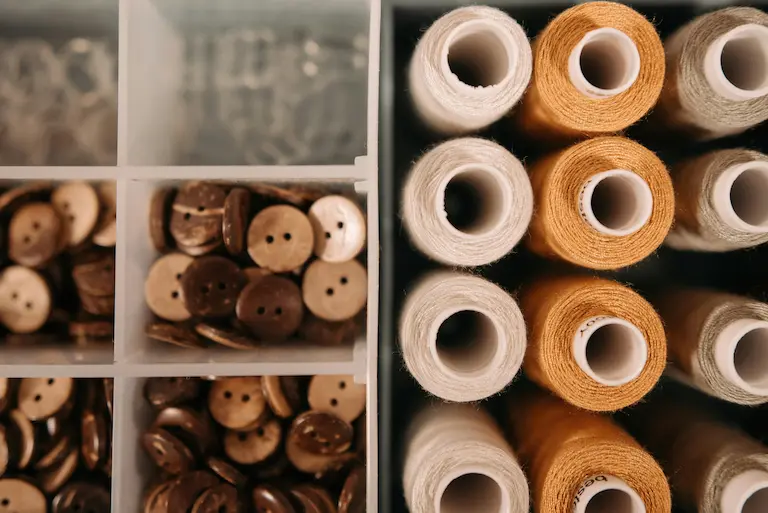 Boîte de couture avec bobines de fil (blanc, beige, gris) et compartiments remplis de boutons en bois.