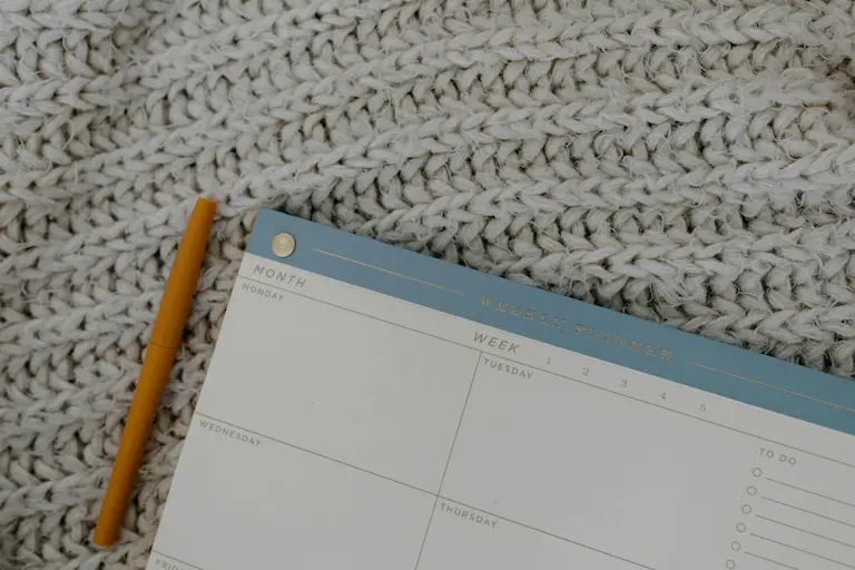 Agenda hebdomadaire posé sur une couverture tricotée, avec un stylo orange à côté.