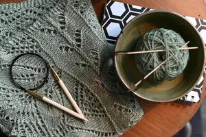 Projet de tricot : châle ajouré vert avec aiguilles circulaires et pelote de laine dans un bol, posé sur une table en bois.