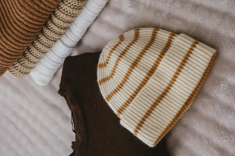 Bonnet en tricot crème à rayures beige, posé sur des vêtements pliés sur une couverture.