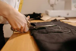 Gros plan sur une main coupant du tissu noir avec des ciseaux sur une table de coupe, machine à coudre floue en arrière-plan.